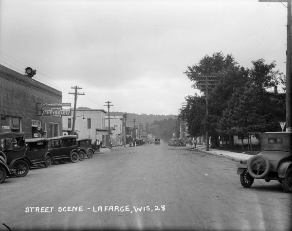 View down a main street with storefronts, showing large power lines stretching into the distance, and a hill in the background. There are cars parked in front of a Chevrolet service station on the left. Pedestrians are on the sidewalk and in the road.