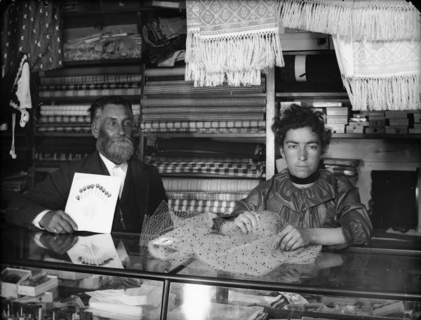 Mr. Meinholtz and a woman, probably his wife or daughter, posing at the counter inside the Meinholtz Store. She is holding a piece of fabric, and he is holding a card of pins.