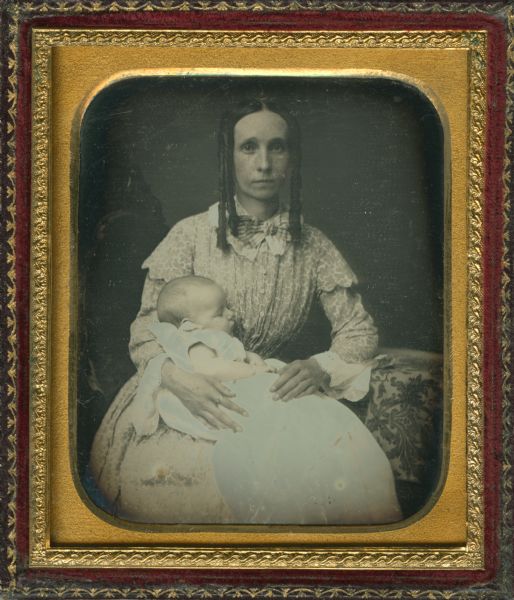 Sixth plate daguerreotype of a seated woman in a long-sleeve printed dress and shoulder-length side curls, holding a sleeping baby on her lap with her right arm. The baby is wearing a long white sleeveless baby gown, tinted blue. Cheeks hand tinted. 