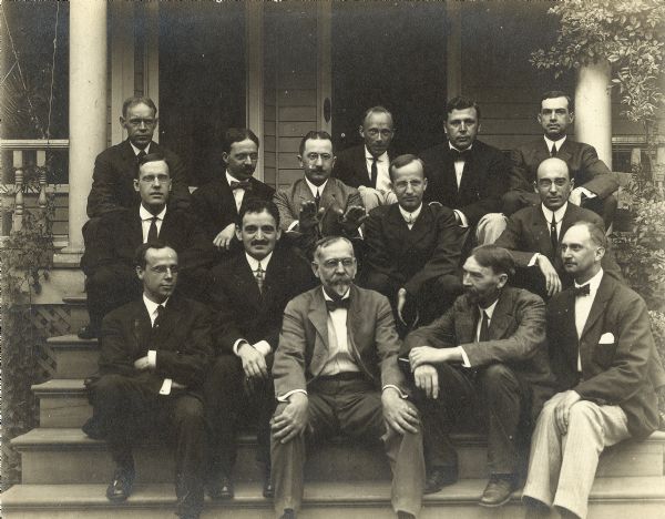 A group portrait of men, possibly economists, seated on the porch stairs in front of a house. The man who sits in the first row, second from the right, is thought to be Thorstein Veblen.