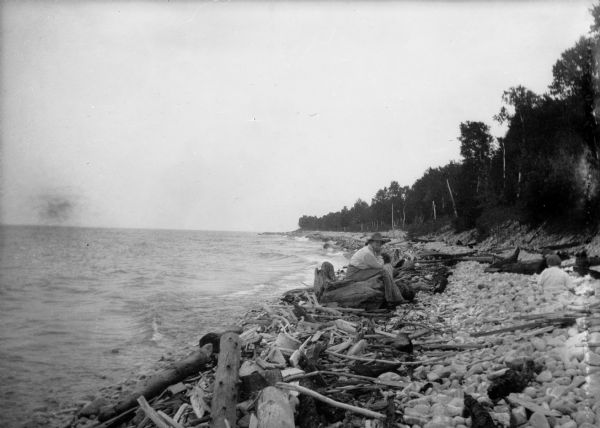 Thorstein Veblen sitting on a beach, probably at Washington Island, surrounded by driftwood. There is a young child on the beach on the right.