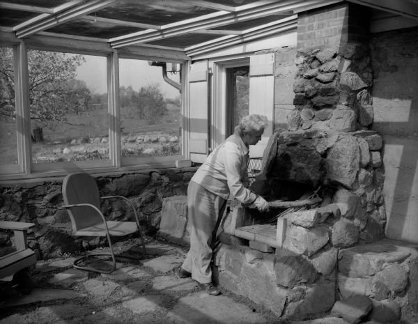 Ruth B. Glassow, a professor of physical education for women at the University of Wisconsin, stacks wood in the outdoor fireplace that she built by herself. The porch was built around the fireplace at a leter date. Her home is at 1635 Norman Way and was a barn on the estate of John R. Commons, a world-renowned University of Wisconsin economist.
