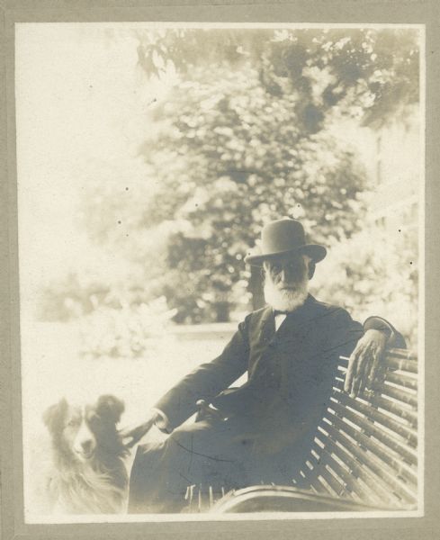 Samuel Marshall is seated on a bench outdoors with his dog Mac at his side. Marshall is wearing a hat and has a white beard.