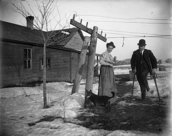 Mr. and Mrs. Jerome J. Moore outdoors at their home next to the Wisconsin Central Railroad tracks. Mr. Moore has a pipe in his mouth and is walking on crutches. A small black dog stands near Mrs. Moore, and snow covers the ground.