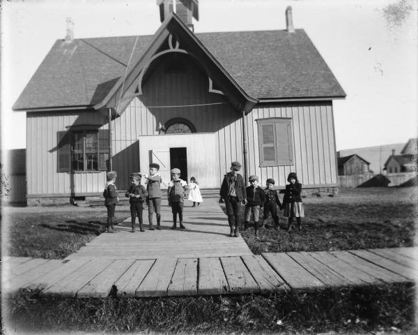 Children posing on a board sidewalk in front of a schoolhouse in New London. The boys are wearing short pants, leggings, and caps, and the girls are wearing skirts and dresses.