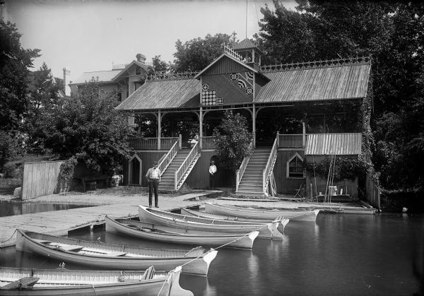 View of the Draper Hall boathouse on Lac La Belle with a row of boats in front of the building. Two men stand on the pier.