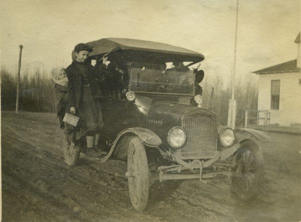 Warmly-dressed school children riding on the running boards of Theta Mead's car. There is a large building and probably a flag pole in the background.
