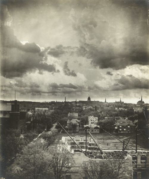 Elevated view over the State Historical Society of Wisconsin building (Wisconsin Historical Society) which is under construction. The Wisconsin State Capitol is silhouetted in the background and the sky is filled with dramatic clouds. The Armory (Red Gym or Old Red) is on the left along Langdon Street.
