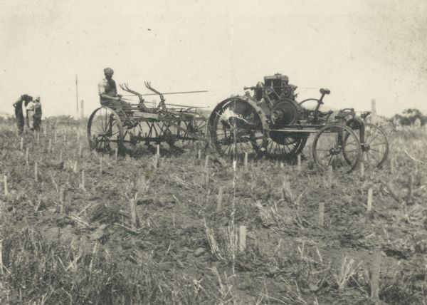 A farmer in a field sits on a cultivator and drives a horseless tractor with reins or belts. Three men are standing in the background.