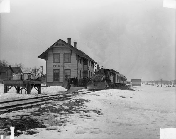 The Illinois Central Railroad Station and train in Fitchburg. There is snow on the ground, and men and women are waiting outside the depot on the platform. There are milk cans on a wooden platform near the tracks, and a windmill in the background near houses.