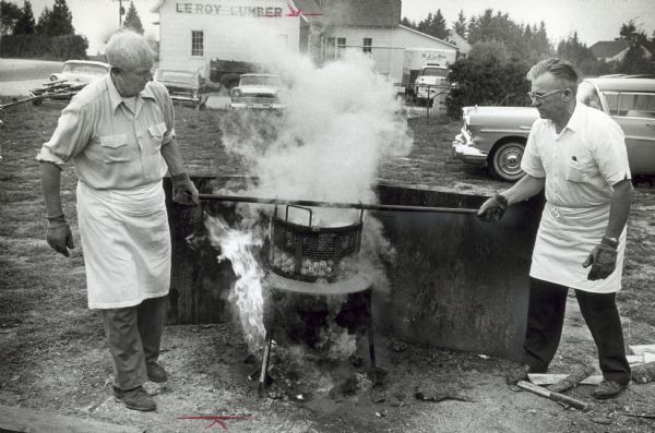 Closely timed cooking and quick serving are the secret of a successful fish boil. A heavy iron bar is inserted in the basket of potatoes and onions to remove it from  the steaming kettle. Lawrence Wickman (right), owner of the Viking helped chef Helmer Erickson. Both men are wearing asbestos gloves.