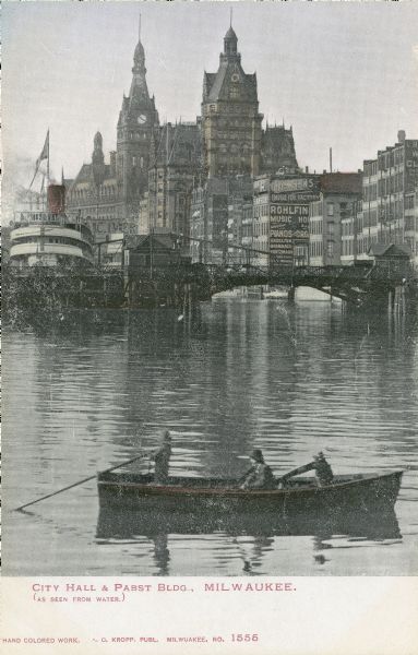 Hand-colored view from the water. Buildings are in the background, a boat and bridge are in the center. A small boat with people in it are in the foreground. Caption reads: "City Hall and Pabst Bldg., Milwaukee. (as seen from water)."