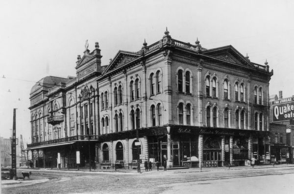 View from street of theatre and main entrance on corner of Wells and Water Streets. To the right is a building with a sign for Quaker Oats. On the left corner is a dog standing in the street.
