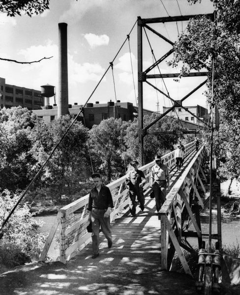 Uniroyal workers cross a suspension bridge over water at the end of a work shift. In the background are factory buildings and a smokestack.