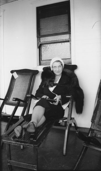 A fashionably dressed woman with a fur stole around her shoulders relaxes on a lounge chair.