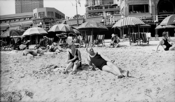 Mr. and Mrs. Brandel relax on a beach, wearing bathing suits. Chairs and umbrellas are behind them, and in the background is a balcony or boardwalk, with shops, and hotels.