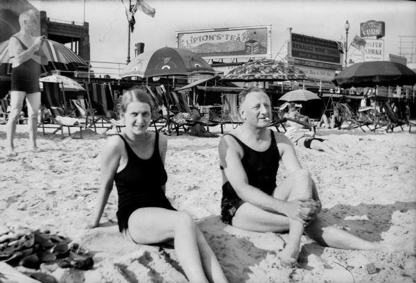 Mary Brandel and Edward (E.W.) Brandel wearing bathing suits relax on a beach during a vacation. Chairs and umbrellas are behind them, and in the background is a balcony or boardwalk, with shops, billboards.