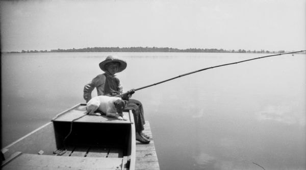 A boy sits in a boat on a pier, with his fishing pole and a dog (Max) at his side. In the background is the far shoreline.