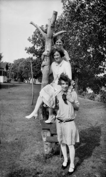 Mrs. Brandel (center) with two other women posing on and near a tree stump on a lawn. There is a building and water in the background.
