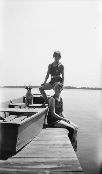 Two young women in bathing suits pose on a pier. Max the dog sits in a small wood boat sitting on the pier. The far shoreline can be seen in the background.