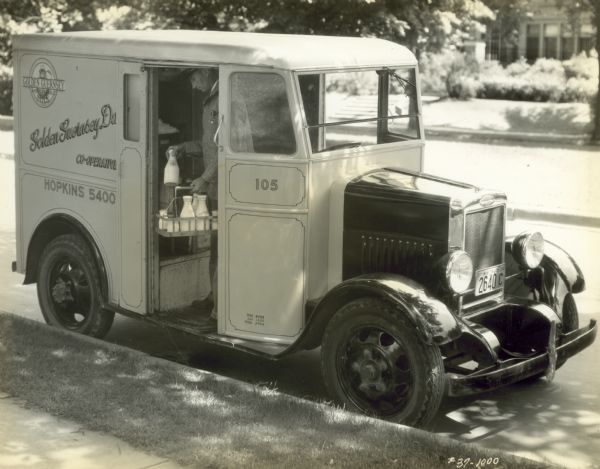 A milkman stands inside a Golden Guernsey Dairy delivery truck loading bottles into a carrier. The truck is parked at a curb and a house is in the background.