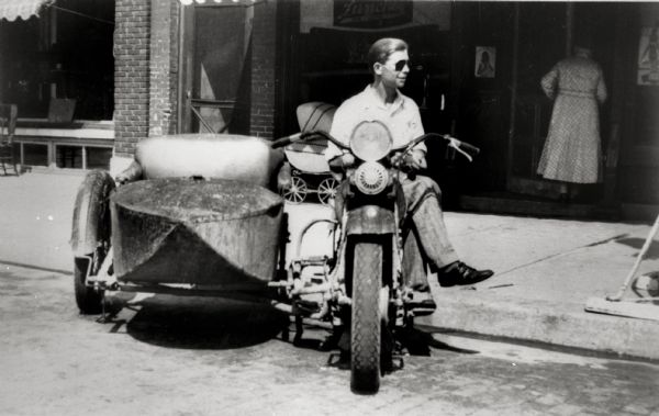 Ralph Hilgers, wearing sunglasses, is seated on his 74 Harley-Davidson twin cylinder, late 1920's model motorcycle. His motorcycle is shown with a prototype of a double-wide sidecar parked in front of The Parkway Cafe, on the corner of Superior Avenue and Juneau Street, owned and operated by Hilger. Ralph's mother Agusta Hilgers can be seen entering the Cafe. Ralph Hilger's son Rollie Hilgers is in a baby carriage parked in front of the restaurant directly behind the motorcycle.