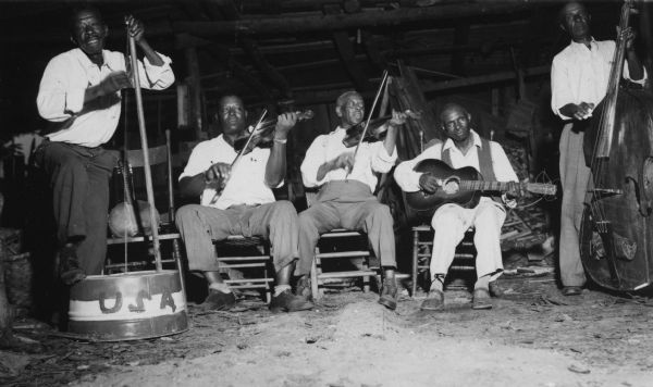 Five African American men with instruments inside a shed or barn: an upside down gutbucket with USA painted on the side, 2 violins, a guitar, and a stand-up bass. The two men on the end are standing, and the three in the middle are sitting on wooden chairs.