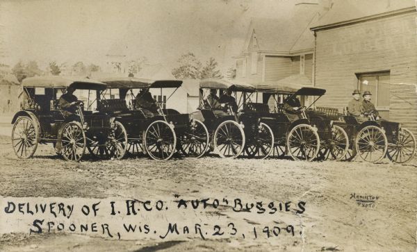 Five International Harvester Auto Buggies with men sitting inside are on a dirt road in front of the Spooner Lumber Company. Caption reads: "Delivery of I.H. Co. 'Auto Buggies,' Spooner, Wis. Mar. 23, 1909."