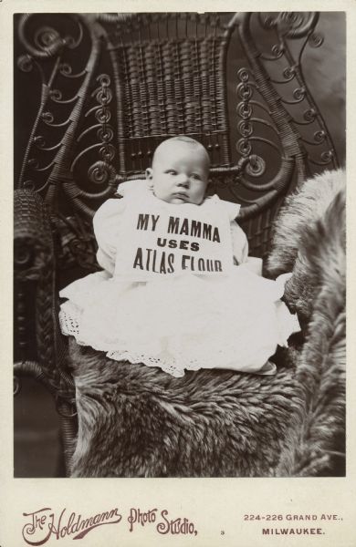 Studio portrait of an infant seated on an animal skin on an elaborately woven wicker chair, wearing a baby dress and a bib reading "my mamma uses Atlas flour."