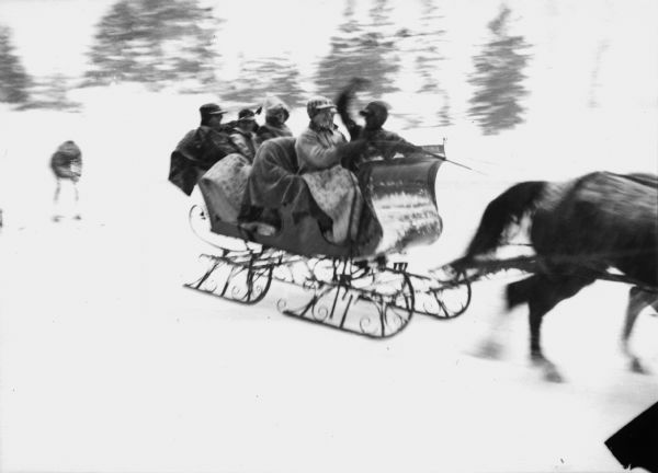 Winter scene with sleigh carrying six well-bundled passengers pulled by two horses and pulling a skier behind in Eagle River. The trees are blurred.