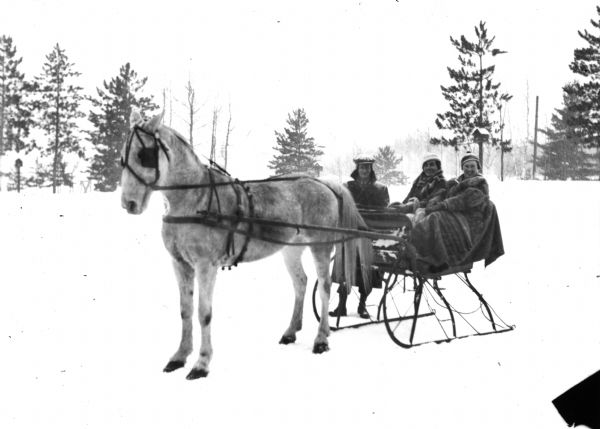 Winter scene with two women wearing hats and coats in a sleigh pulled by a white horse over snow, Eagle River, Wisconsin. A man stands beside the sleigh, and in the background are trees.