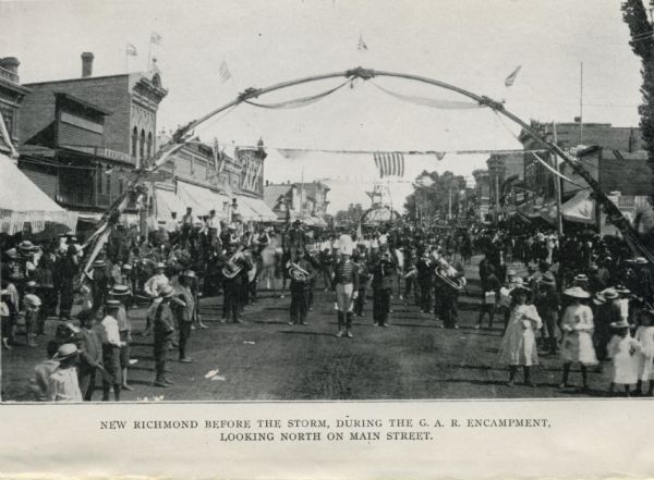 View of a marching band parading beneath a decorated arch on Main Street. Onlookers line both sides of the street. American flags are flying over the road and from rooftops. There is another arch in the background. Original caption reads: "New Richmond before the storm, during the G.A.R. encampment, looking north on Main Street."