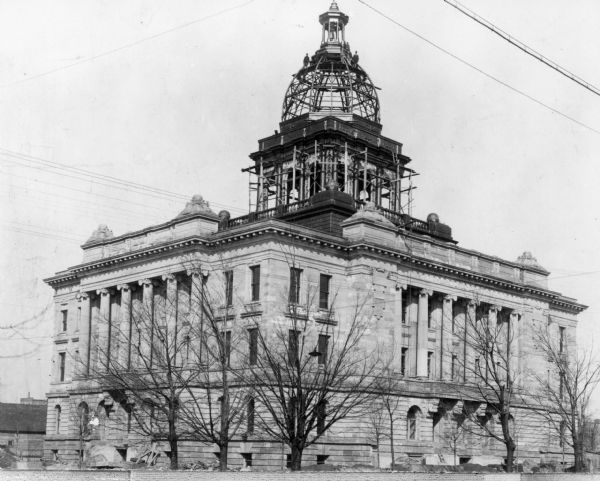 Exterior view of construction on the building. There are gentleman standing on the iron framework of the dome. The dome originally was encased in glass, and later sheet metal after a disastrous hailstorm broke a number of the glass panes. The Classical styling of the nearly completed courthouse was typical of the time period. 

