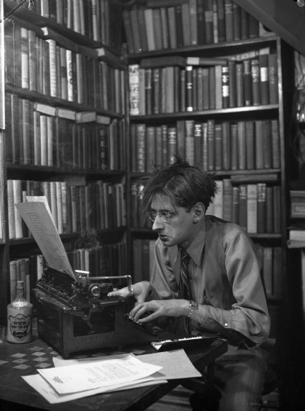 Robert Albert Bloch, author of <i>Psycho</i>, surrounded by tall bookshelves full of books, is sitting in front of a typewriter with an open cigarette case full of cigarettes, a bottle of Wing Fhung Hong, and a pile of papers.