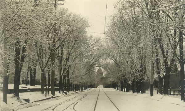 Winter scene up the center of a tree-lined State Street from the 600 block, with the Wisconsin State Capitol visible in the distance. Streetcar tracks are visible in the snow.