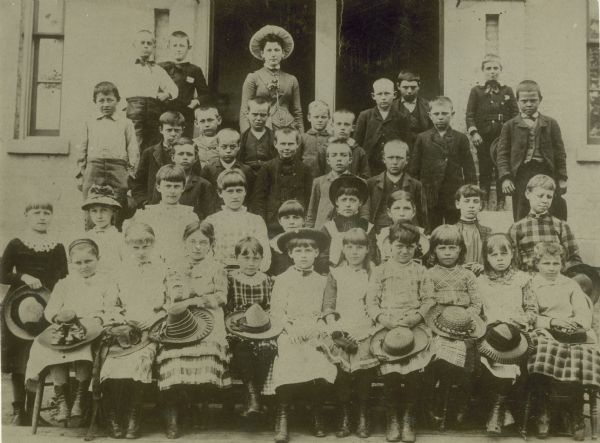 A group portrait of students at the 2nd Ward School. The students are posing in front of the building, with girls in the front (straw hats on laps), boys in the back, and their teacher is in the back in the middle. On the back of the photograph, in what appears to be a child's handwriting, are the following names (as they are spelled by the writer): Miss Eliza Herfuth, Sidney Ainsworth, Hary Bancroft, Arthur Baker, John Lavery, Bertrand Doyon, Eddie Swain, Clay Beryman, John Nebhour, Harry Deards, Robie Ryder, George Cain, Hary Stollze, Jimmie Livesy, and John Port. There is no indication of which children in the photograph these names apply to.