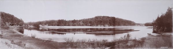 Panoramic view from shoreline of the Wisconsin River in the Wisconsin Dells as seen from the "Old Dell House." A canoe is visible on the shore on the far right, and behind it is a covered excursion boat. In the lower right corner on a label are the words: "THE WISCONSIN DELLS, FROM THE OLD DELL HOUSE. Copyright 1894. by H.H. Bennett."