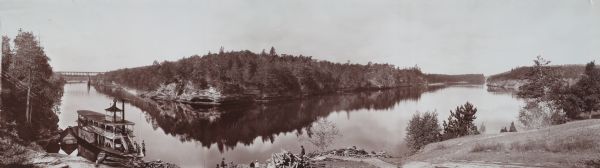 Elevated panoramic view of the Wisconsin River shoreline in the Wisconsin Dells. There is an excursion boat, <i>The Dell Queen</i>, visible in the lower left corner. People are visible on the boat as well as on what appears to be a dock on the shore.