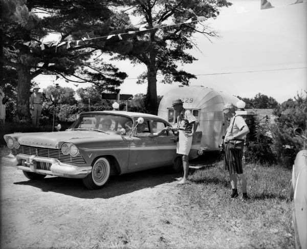 Two men with cameras around their necks stand near a couple in a Plymouth pulling an airstream camper through a gate. The sign on the gate in the background says: "Chula-Vista Farm."