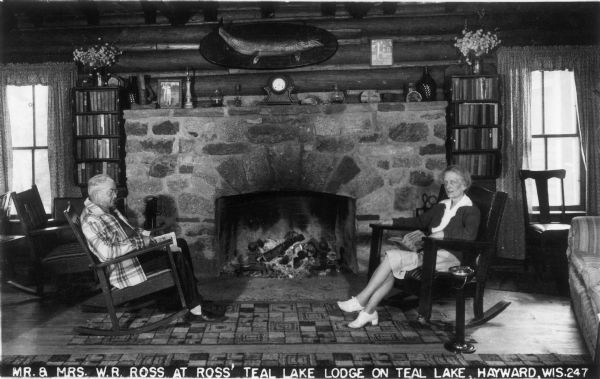 Walter and Virginia Ross relaxing indoors by the fireplace at Ross Teal Lake Lodge. Caption reads: "Mr. & Mrs. W.R. Ross at Ross' Teal Lake Lodge on Teal Lake, Hayward, Wis." 