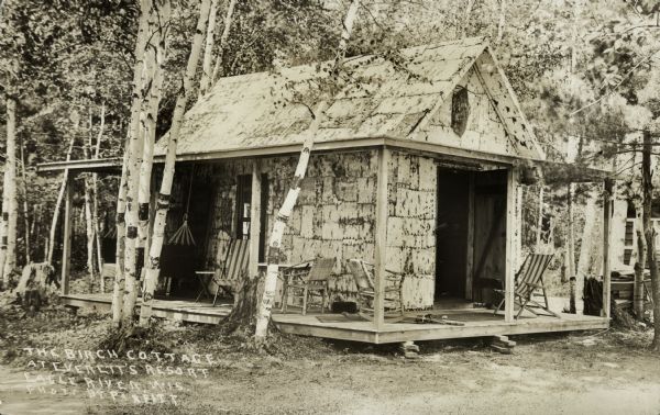 Exterior view of the Birch Cottage surrounded by trees. There is a cushioned porch swing and other outdoor furniture on the rustic porch which wraps around the structure. Caption reads: "The Birch Cottage at Everett's Resort, Eagle River, Wis."