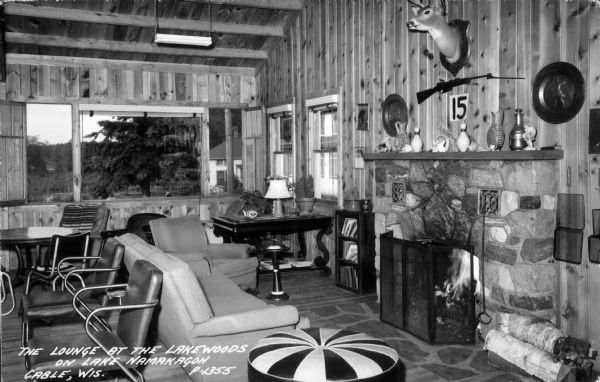 The lounge at the Lakewoods on Lake Namakagon, which includes a fireplace, furniture, large picture window, and a mounted deer head on the wall. Caption reads: "The Lounge at the Lakewoods on Lake Namakagan, Cable, Wis."