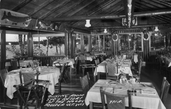 View of the dining room which shows tables set for a meal. Large picture windows have views of the lake and woods surrounding the resort, and above the windows are a number of mounted fish and  two deer heads. Caption reads: "Dining Room at Syd's Resort of the woods, Boulder Jct. Wis."