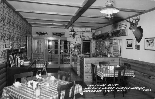 Interior view of the trading post at Willie's White Birch Lodge. Fishing lures are on display on the back wall behind a counter with signs that say: "Pat's Swap Shop," "Open for Trading," and "Trade at Your Own Risk." There are tables with gingham tablecloths in the foreground, and examples of taxidermy on the walls.