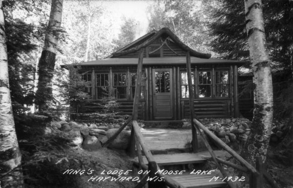 Exterior view of walkway with a rustic gate leading up to the front entrance of a log cabin. Caption reads: "King's Lodge on Moose Lake, Hayward, Wis."