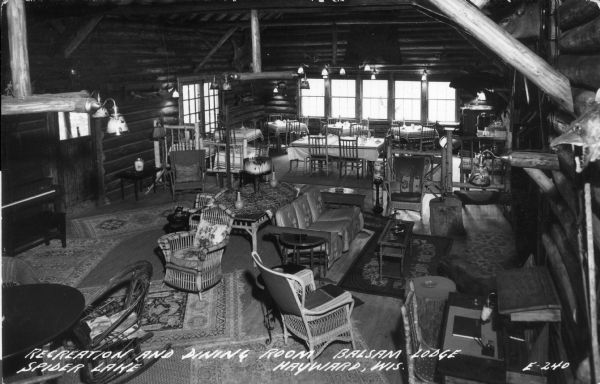 Elevated view of a recreation and dining room. There is a piano near the door on the left, couches, chairs and tables in the foreground, and in the background near a bank of windows, a dining area with chairs and tables. Caption reads: "Recreation and Dining Room, Balsam Lodge, Spider Lake, Hayward, Wis."