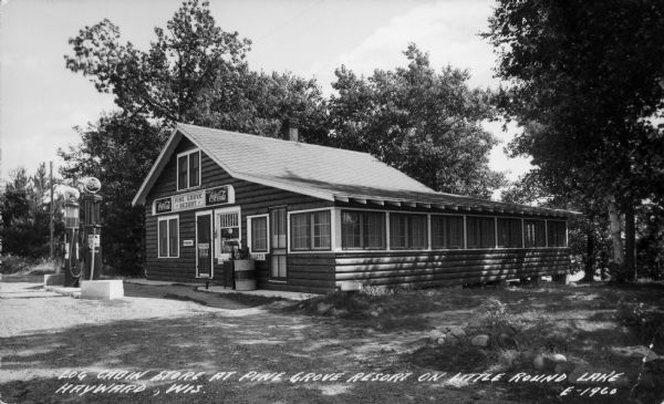 Exterior view toward the store. There are two gas pumps in front, and trees and a lake in the background. Caption reads: "Log cabin store at Pine Grove Resort on Little Round Lake, Hayward, Wis."