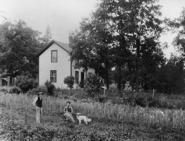 View of Oak Hill Farm, including the farmhouse. In the foreground are a man standing on the left, a woman sitting with a cat in her lap, and a dog.