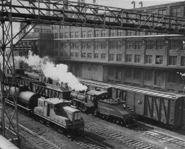 Elevated view of Allis-Chalmers plant showing several trains on the tracks next to a large industrial building. The steam engine tender in front of the locomotive has written on its side: "Allis-Chalmers Mfg. Co."