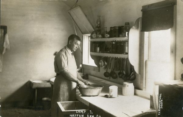 A man works in a kitchen at Hatton Lumber Company Camp. He is wearing an apron and is mixing ingredients in a large bowl at a counter near two windows. Caption reads: "Camp 3, Hatton Lbr. Co., Wis."